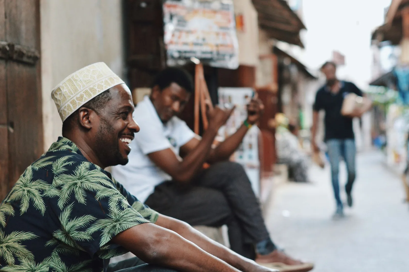 Deux habitants de Stone Town, à Zanzibar, bavardent et rient assis dans une ruelle, l'un coiffé du kofia traditionnel